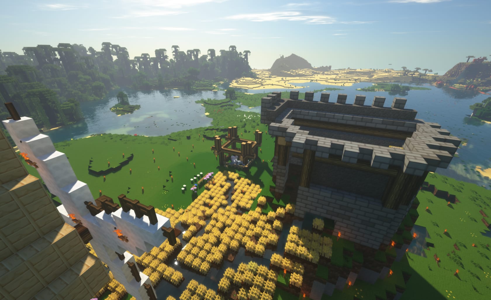 Screenshot of Minecraft game (Using PureBDCraft Minecraft Resource pack https://bdcraft.net) depicting: Grassy landscape with a windmill in the bottom left with a building center right both on a hill with a field of wheat below.