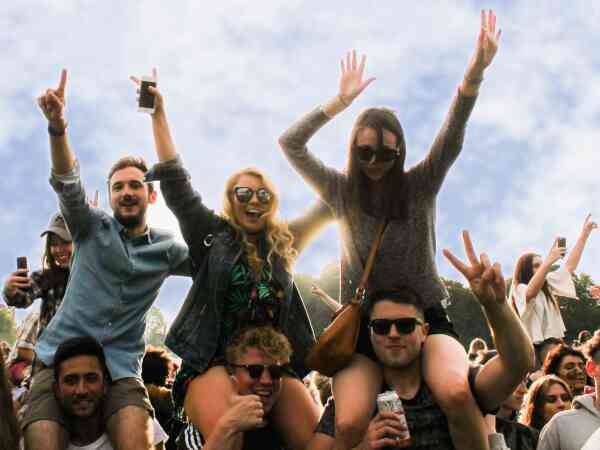 People wearing sunglasses with their arms in the air smiling with some sitting on the shoulders of those underneath.